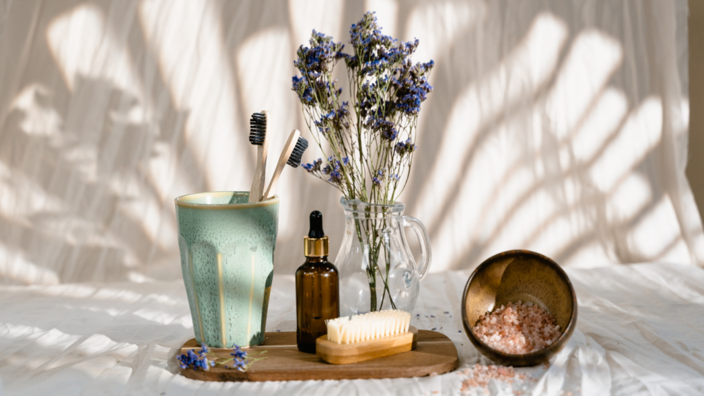 bodegón con un vaso verde y cepillos de dientes de bambú, sobre una bandeja de madera junto a un jarrón transparente con flores
